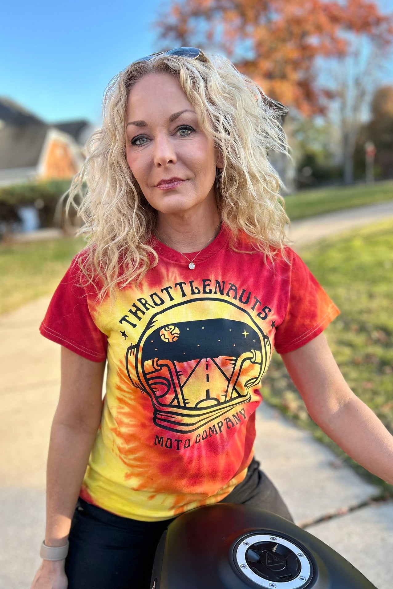 Woman wearing a tie-dye t-shirt with a motorcycle graphic and text, sitting on a motorcycle.